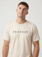 A man wearing a light beige crewneck t-shirt with 'SALVATION' printed in black across the chest, photographed against a neutral light gray background.