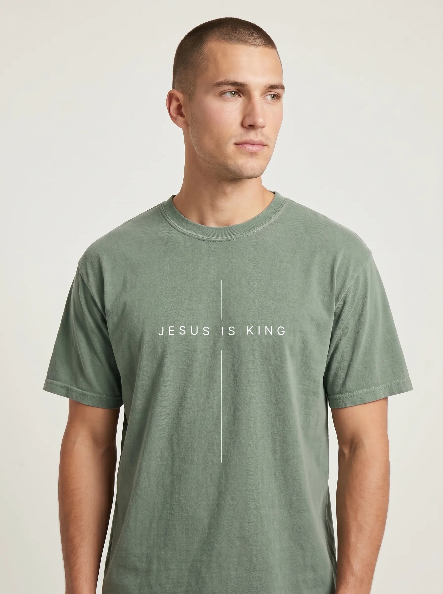 A man wearing a muted sage green crewneck t-shirt with the white printed text 'JESUS IS KING' and a small cross on the chest, photographed against a light gray studio background.