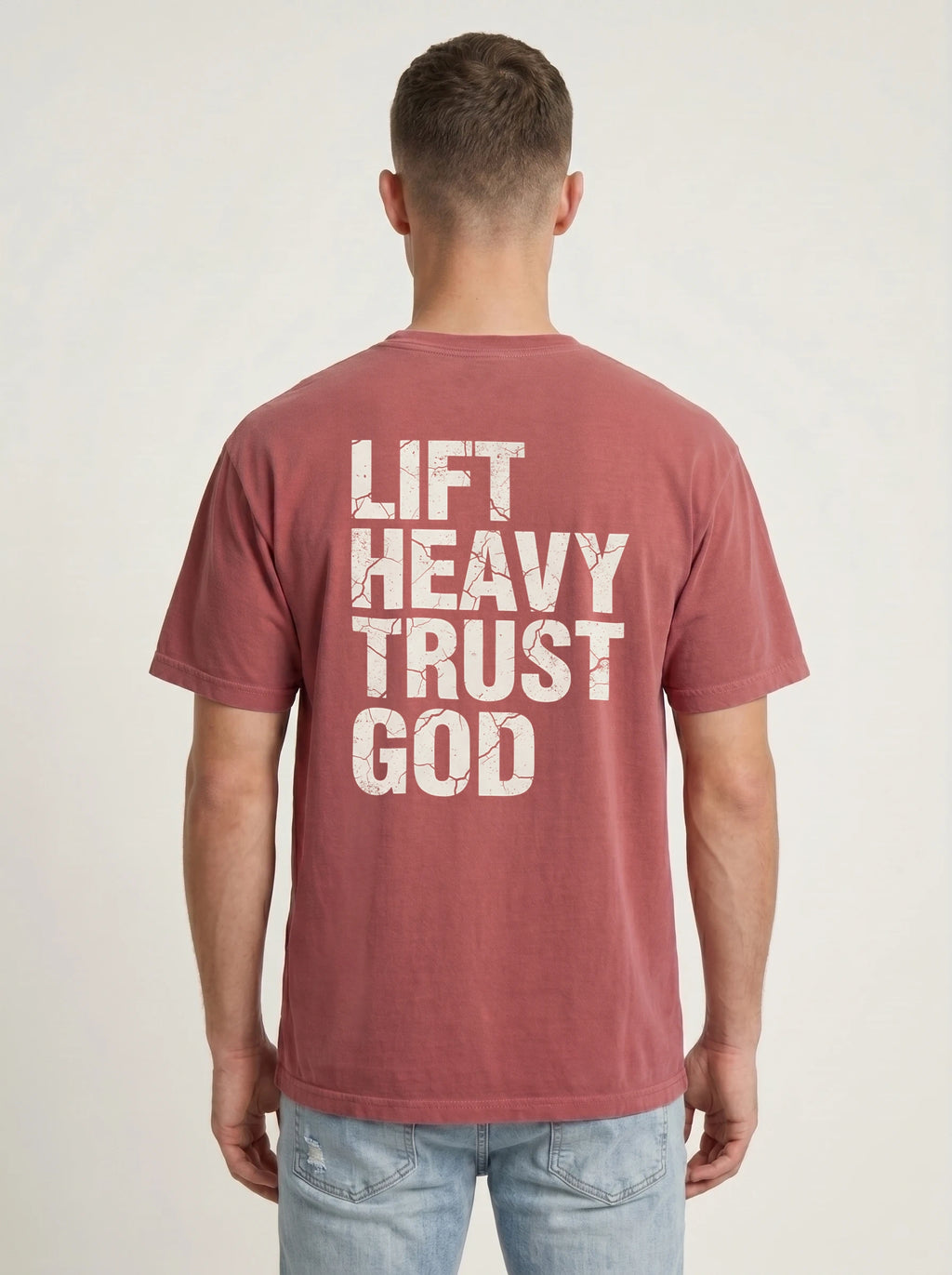 A man viewed from behind wearing a faded dusty rose t-shirt with the text 'LIFT HEAVY TRUST GOD' printed in distressed white block letters on the back, paired with light blue jeans against a light gray studio background.