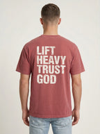 A man viewed from behind wearing a faded dusty rose t-shirt with the text 'LIFT HEAVY TRUST GOD' printed in distressed white block letters on the back, paired with light blue jeans against a light gray studio background.