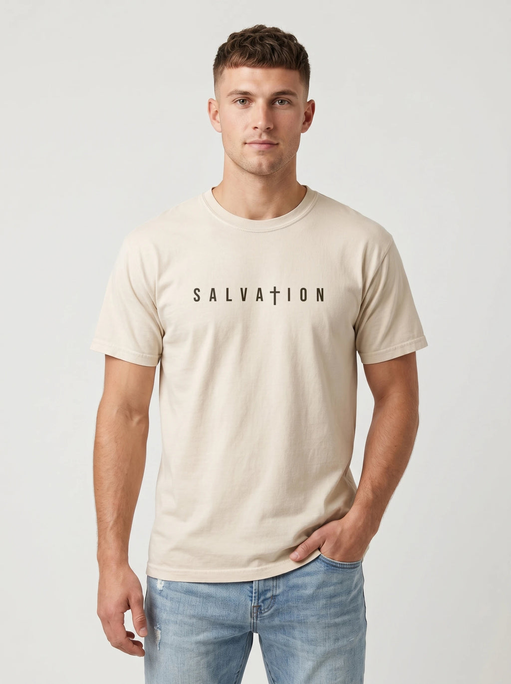 A man wearing a light beige crewneck t-shirt with the word 'SALVATION' printed in black across the chest, paired with light blue jeans, against a plain light gray background.