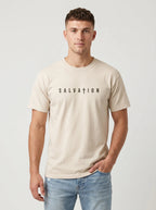 A man wearing a light beige crewneck t-shirt with the word 'SALVATION' printed in black across the chest, paired with light blue jeans, against a plain light gray background.