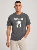 A man wearing a dark gray graphic t-shirt with a white Spartan helmet and the word 'SALVATION' printed on the chest, paired with beige pants, against a light gray studio background.