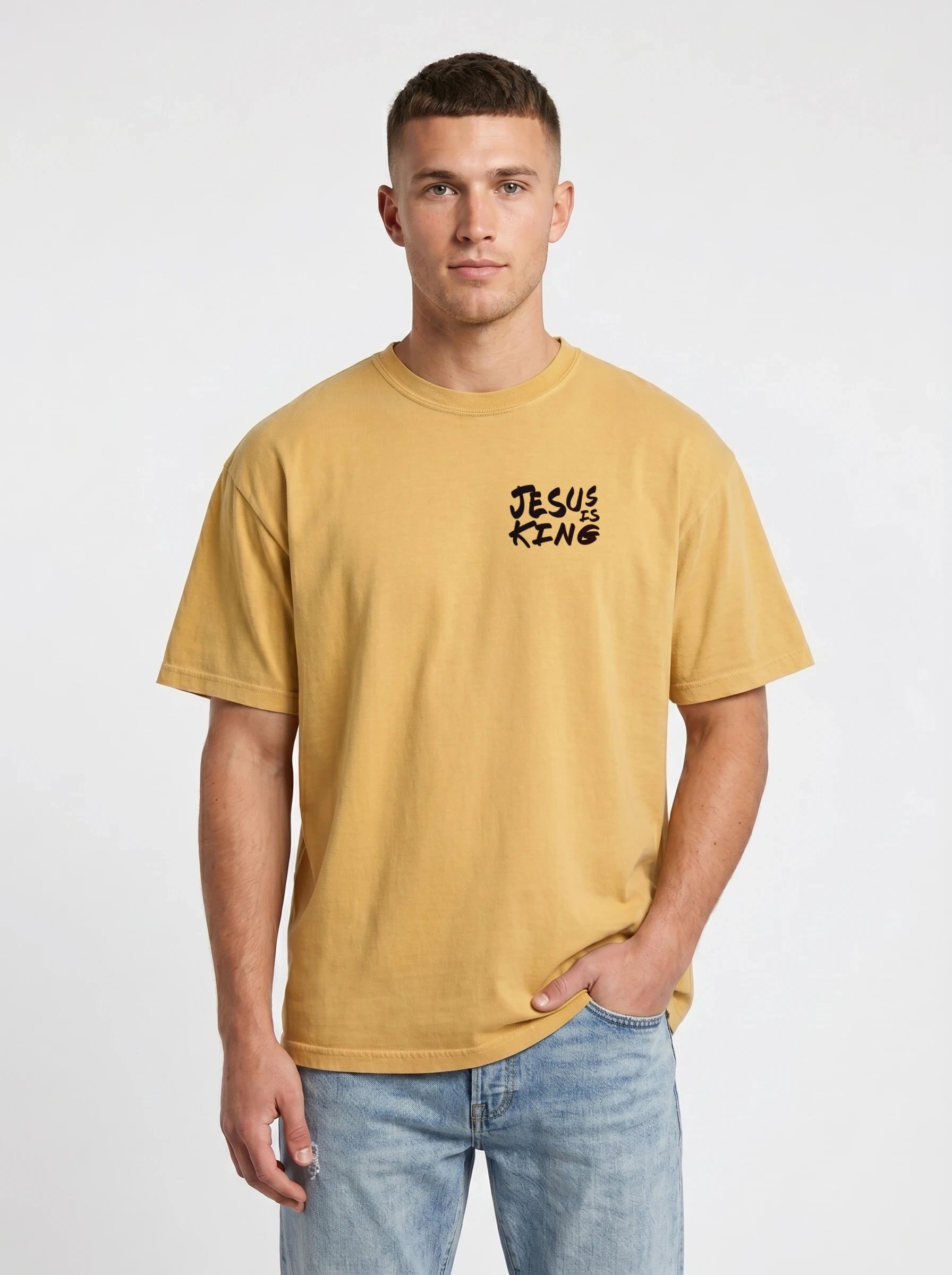 A man wearing a mustard yellow t-shirt with a black 'JESUS KING' graphic on the left chest, paired with light blue jeans, against a plain light gray background.