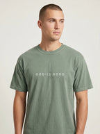 A man wearing a muted sage green crewneck t-shirt with the white printed text 'GOD IS GOOD' on the chest, photographed against a neutral light gray background.
