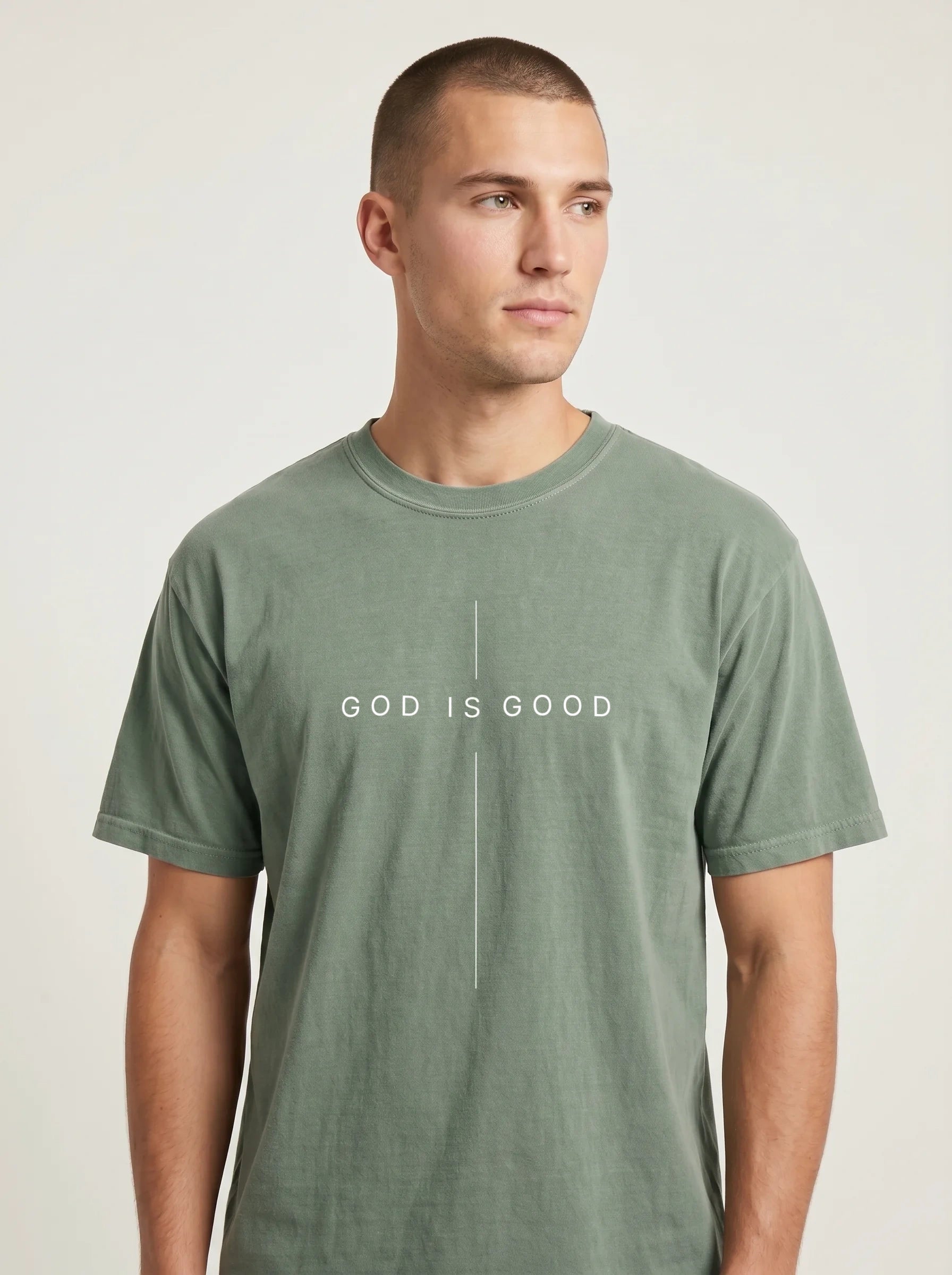 A man wearing a muted sage green crewneck t-shirt with the white printed text 'GOD IS GOOD' on the chest, photographed against a neutral light gray background.