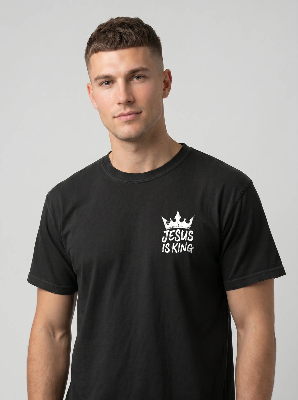 A man wearing a black crewneck t-shirt with a white graphic on the left chest that reads 'JESUS IS KING' beneath a small crown illustration, set against a plain light gray background.