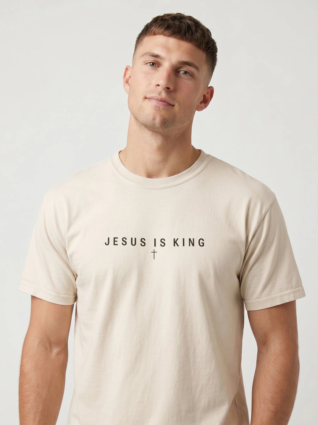 A man wearing a light beige crewneck t-shirt with the text 'JESUS IS KING' and a small cross printed on the chest, set against a plain light gray background.
