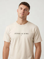 A man wearing a light beige crewneck t-shirt with the text 'JESUS IS KING' and a small cross printed on the chest, set against a plain light gray background.