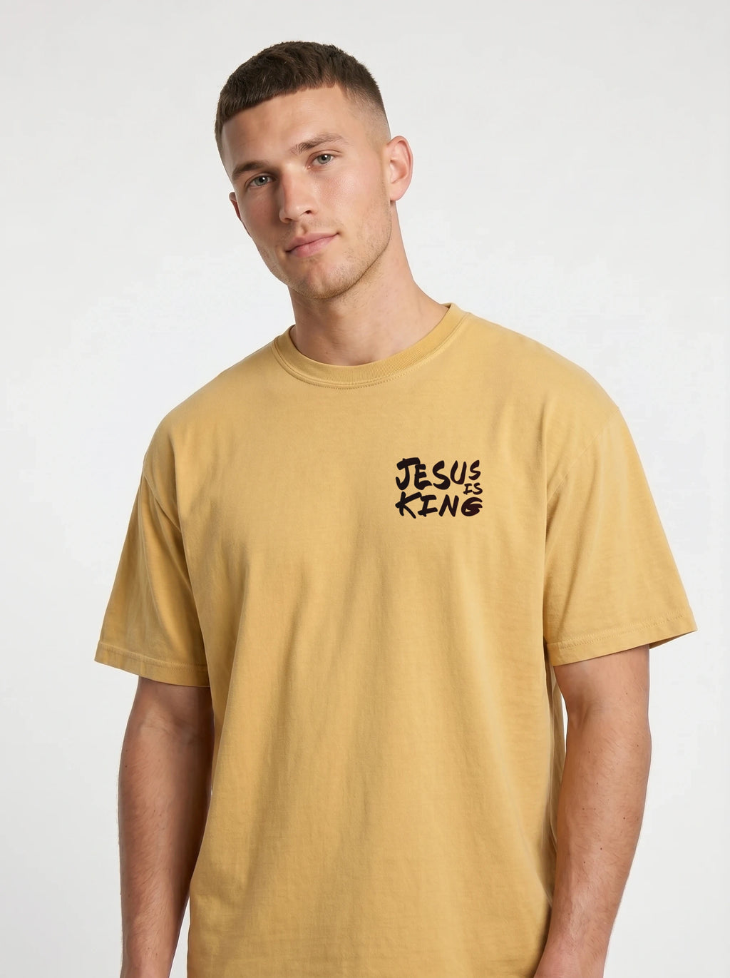 A man wearing a mustard yellow t-shirt with a black graphic text print on the left chest that reads 'JESUS IS KING'.