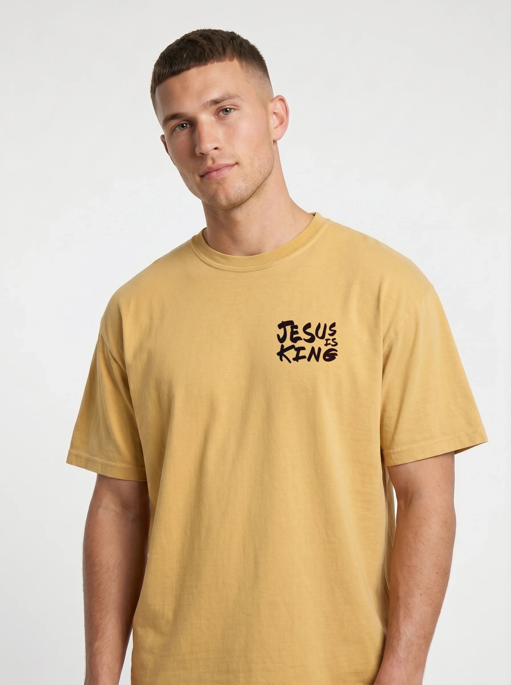 A man wearing a mustard yellow t-shirt with a black graphic text print on the left chest that reads 'JESUS IS KING'.