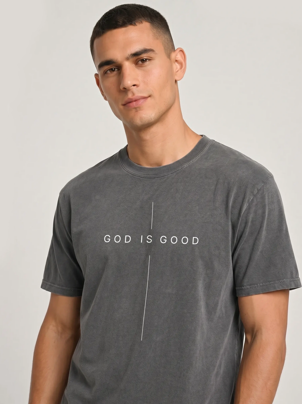 A man wearing a dark gray mineral-wash t-shirt with the text 'GOD IS GOOD' printed in white across the chest, separated by a vertical line.