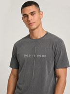A man wearing a dark gray mineral-wash t-shirt with the text 'GOD IS GOOD' printed in white across the chest, separated by a vertical line.