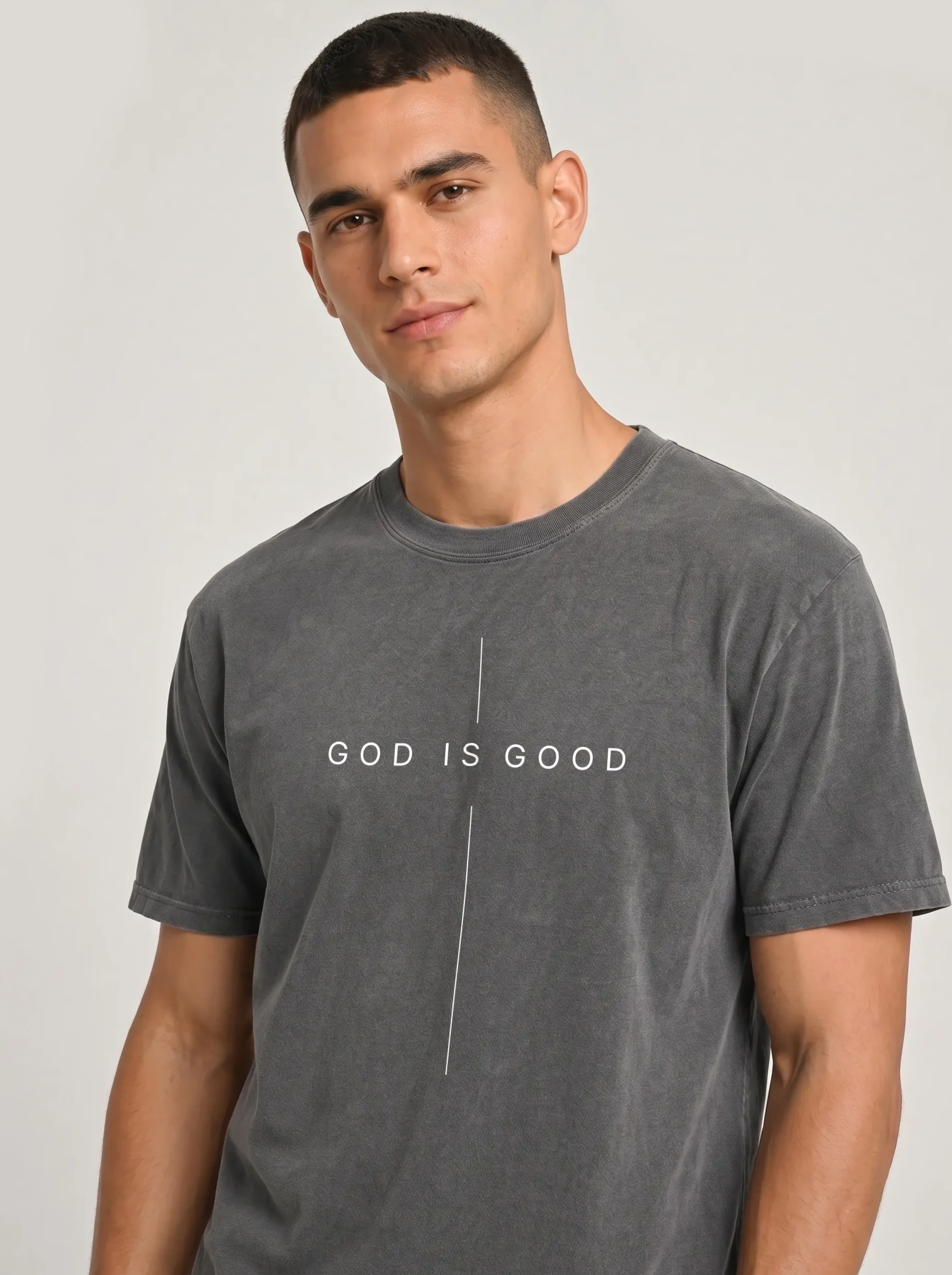 A man wearing a dark gray mineral-wash t-shirt with the text 'GOD IS GOOD' printed in white across the chest, separated by a vertical line.