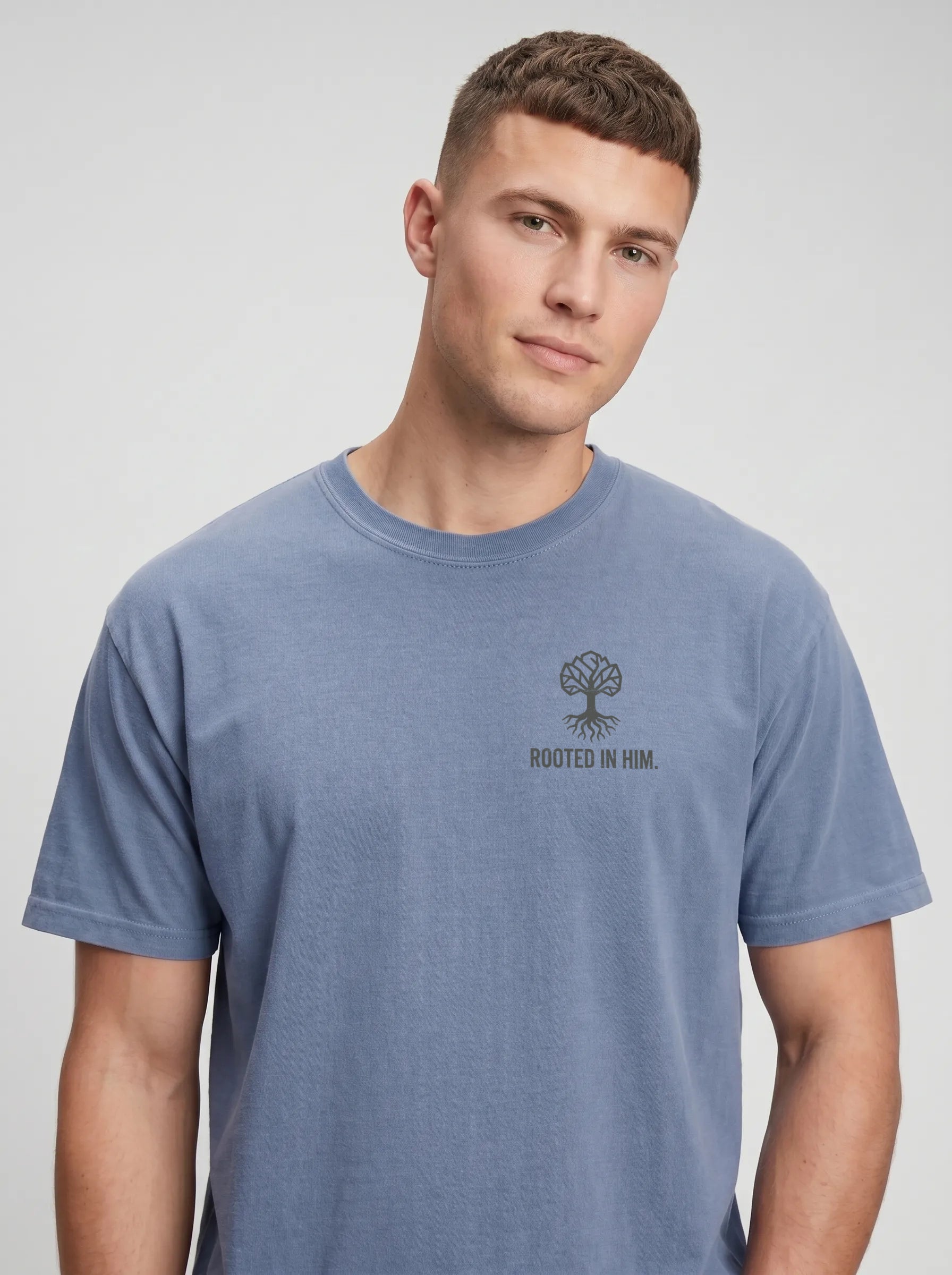 A man wearing a heathered blue crewneck t-shirt with a dark gray tree graphic and the text 'ROOTED IN HIM.' printed on the left chest, set against a plain light gray background.