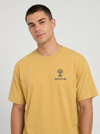A man wearing a mustard yellow t-shirt with a small black tree logo and the text 'ROOTED IN HIM.' on the left chest, photographed against a light gray studio background.