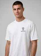 A man wearing a white short-sleeve crewneck t-shirt with a small black chest graphic that reads 'ROOTED IN HIM' beneath a tree logo, set against a plain gray studio background.
