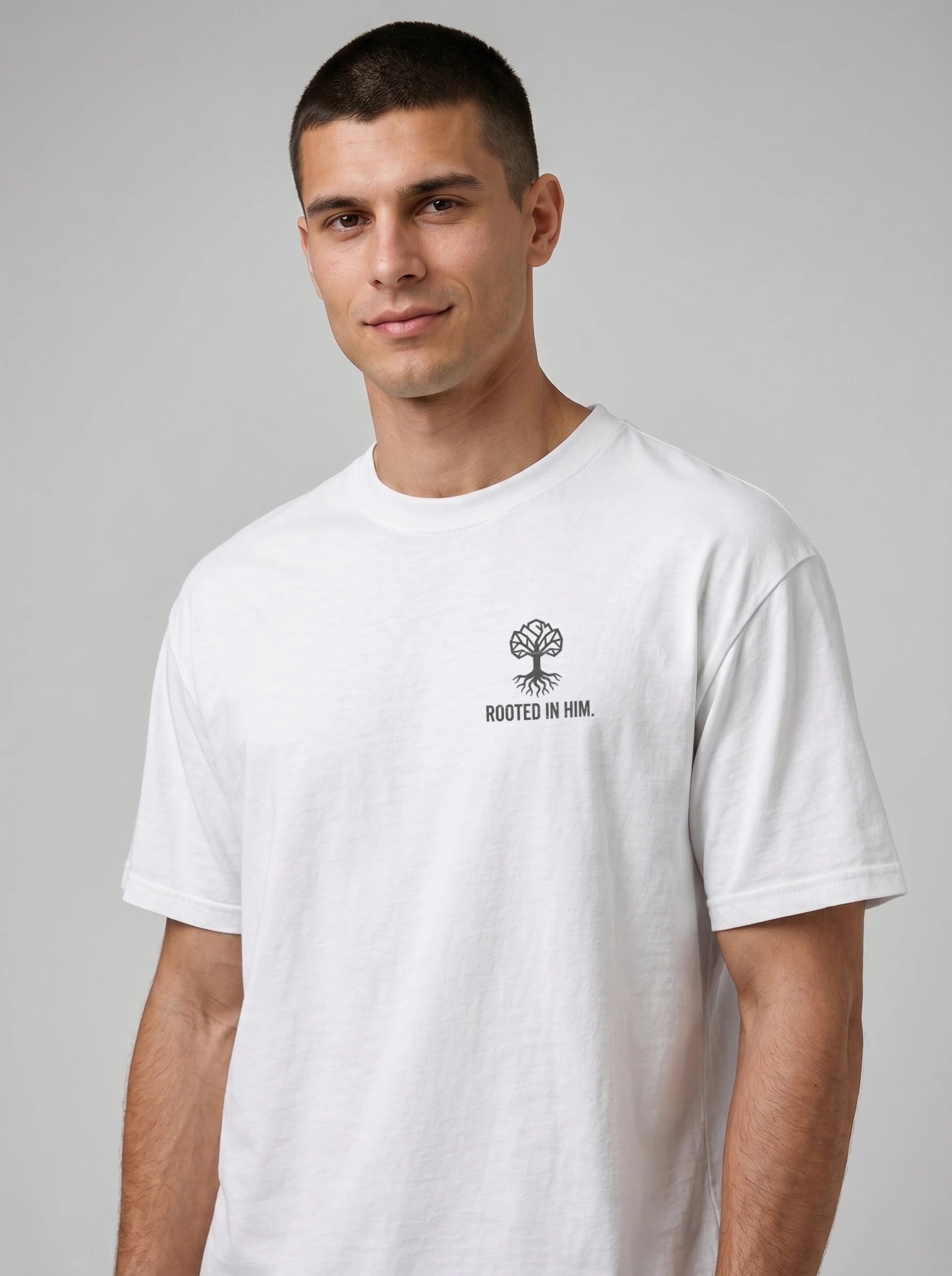 A man wearing a white short-sleeve crewneck t-shirt with a small black chest graphic that reads 'ROOTED IN HIM' beneath a tree logo, set against a plain gray studio background.