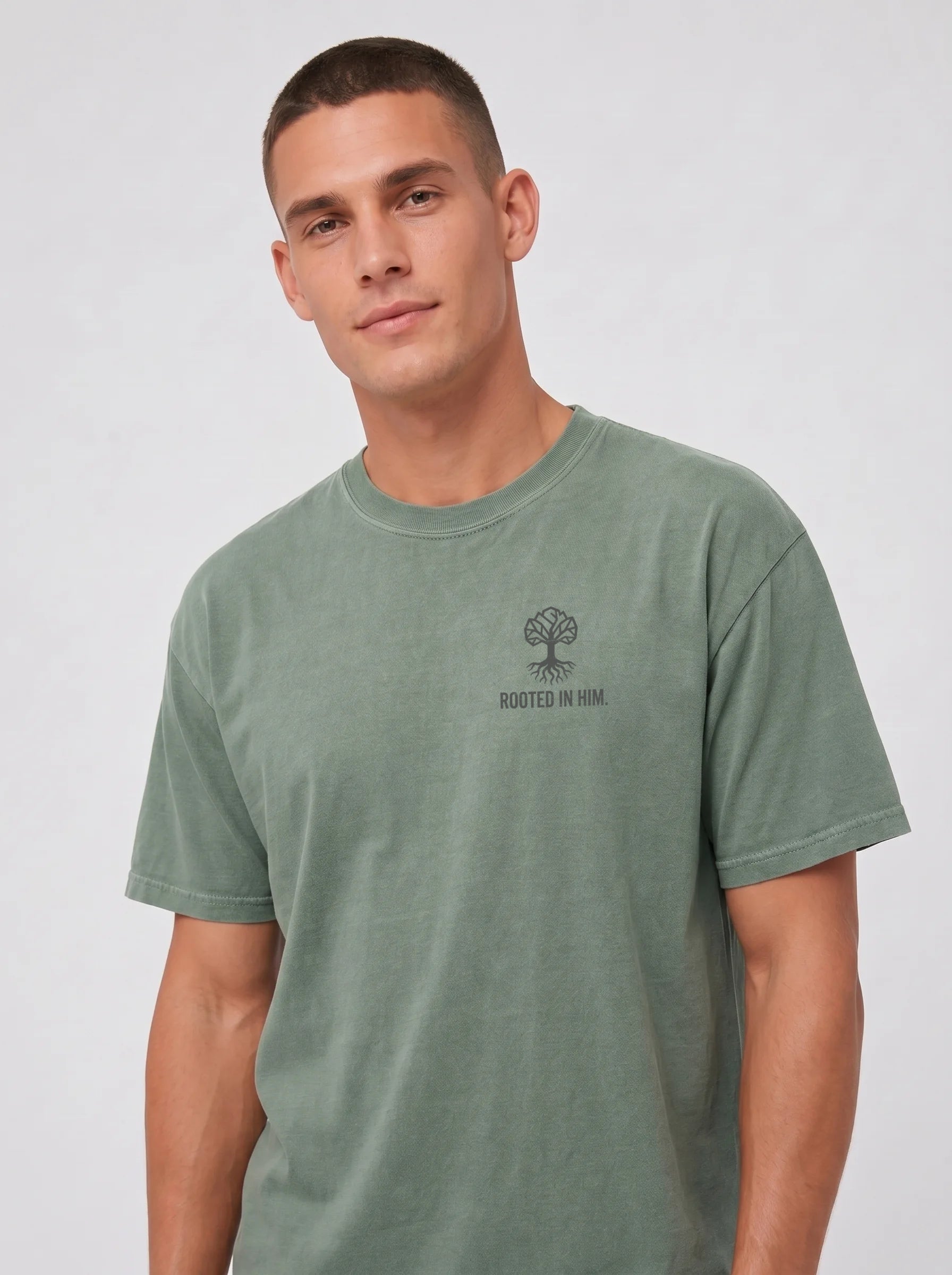 A man wearing a sage green crewneck t-shirt with a small dark gray 'ROOTED IN HIM' tree graphic on the left chest, photographed against a light gray studio background.