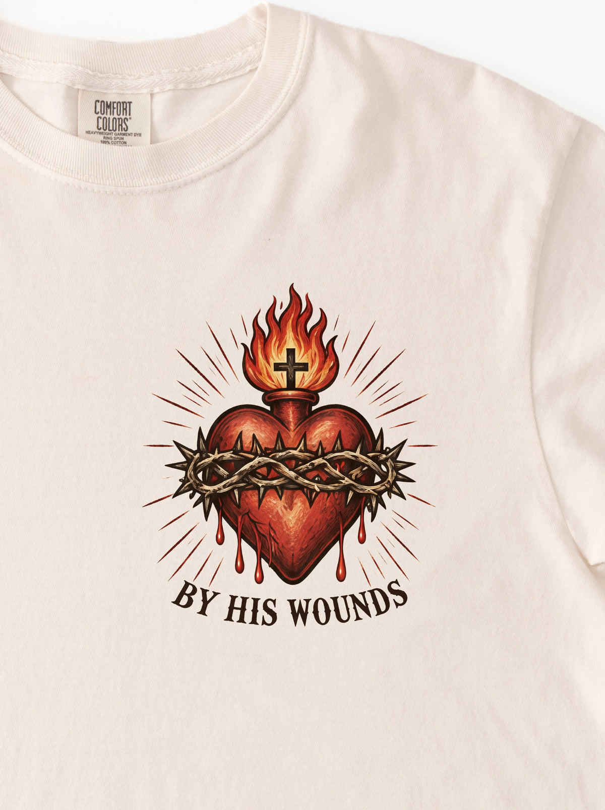 Cream T-shirt with a flaming sacred heart, cross and crown of thorns, with the text “By His Wounds.”
